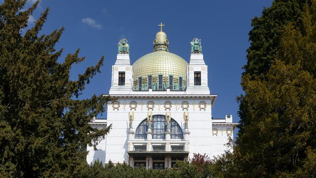 Otto Wagner Kirche am Steinhof, Außenansicht. Zwischen zwei Bäumen strahlt ein weißer Kirchenbau mit einer gold-grünen Kuppel