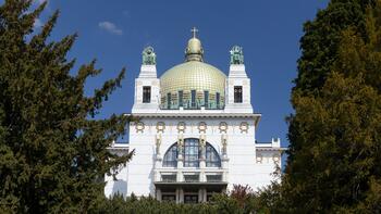 Otto Wagner Kirche am Steinhof, Außenansicht. Zwischen zwei Bäumen strahlt ein weißer Kirchenbau mit einer gold-grünen Kuppel