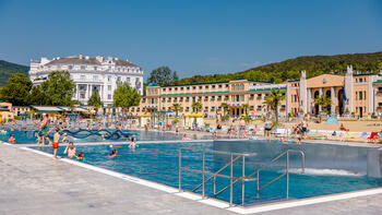 Blick auf die Becken mit Menschen in Badekleidung; dahinter Sandstrand und das Hotel Esplanade; Farbfoto.
