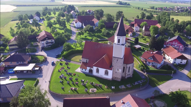 Das Luftbild zeigt die auf einem Hügel errichtete gotische Pfarrkirche des Dorfweilers Münsteuer inmitten der weiten Ebene des unteren Inntals.