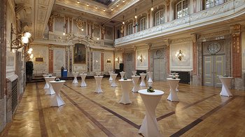 Blick in den großen Festsaal mit Stehtischen und Sprecherpult am Podium. Gut erkennbar die Galerie und das große Portrait von Kaiser Franz Josef.