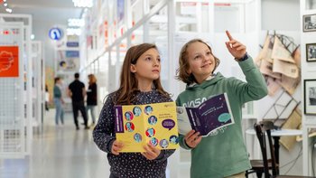 Zwei Kinder mit Rätselheften in einer hellen Ausstellung. Im Hintergrund sind Personen zu sehen.