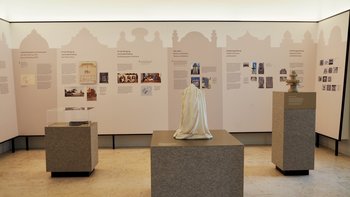 Im Vordergrund ein Podest mit einer verhüllten Vase aus Stein, im Hintergrund Tafeln der Ausstellung zum jüdischen Friedhof Währing.