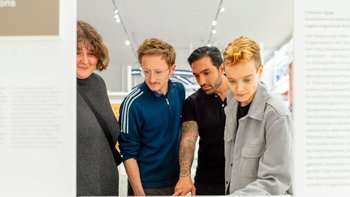 Vier Personen in gedeckten Farben gekleidet, sehen sich Inhalte in einer hellgestalteten Ausstellung an