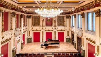 Blick von der Galerie des Ehrbar Saals auf dessen Bühne. Der Saal ist rot und golden ornamentiert, auf der Bühne steht ein schwarzes Klavier.